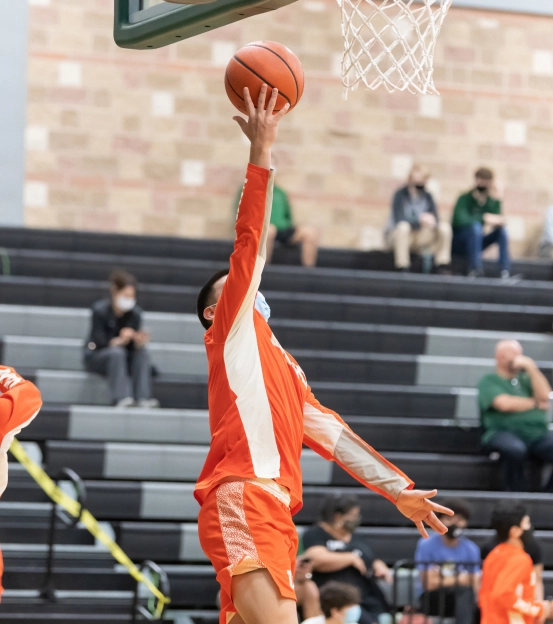 Men's basketball game. A young man in an orange jersey jumps towards the net.